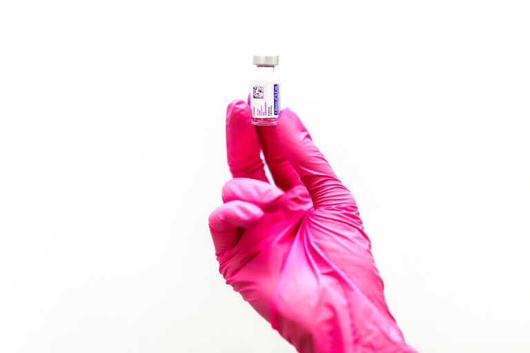 A gloved hand holds a small vial containing a clear liquid against a white background.