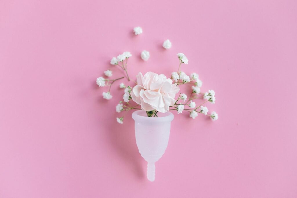 A menstrual cup holding white and pink flowers is placed on a pink background, with small white flowers arranged around it.