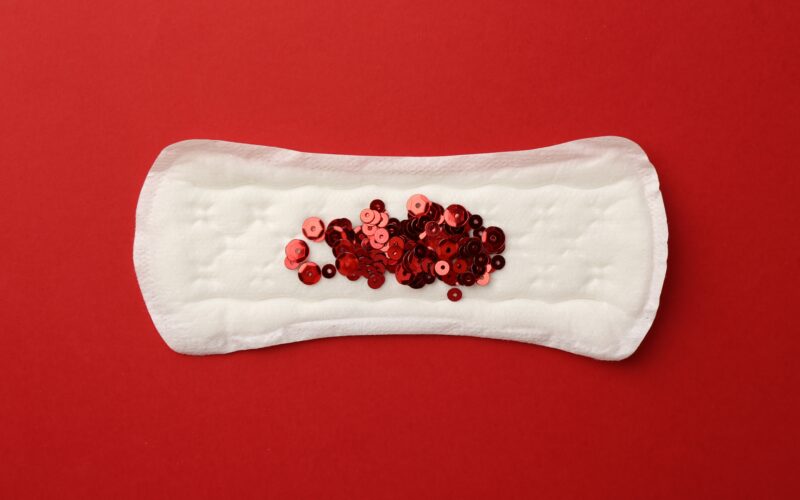 A sanitary pad on a red background with red sequins arranged in the center, resembling menstrual blood.