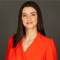 Une femme aux cheveux bruns lisses, portant un chemisier orange vif, pose sur un fond uni et sombre, regardant l'appareil photo et souriant légèrement.